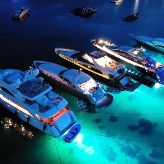 Four luxury yachts anchored at night with bright blue and white underwater and interior lights, illuminating the clear, dark water.